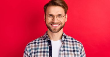 Photo of pretty charming young guy wear checkered shirt glasses smiling isolated red color background.