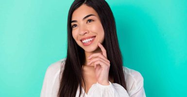 Portrait of pretty minded vietnamese girl finger touch chin look empty space toothy smile isolated on emerald color background.