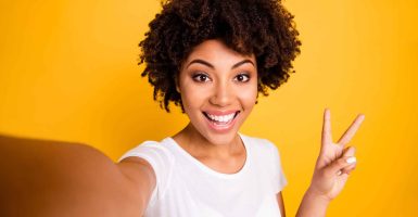 Close up photo beautiful amazing she her dark skin lady arm hand show v-sign make take selfies cute attractive white teeth wear casual white t-shirt isolated yellow bright vibrant vivid background.